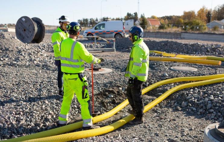 Tre byggarbetare i gula varselkläder och hjälmar står på en grusig arbetsplats bredvid stora gula rör, med en servicebil i bakgrunden.